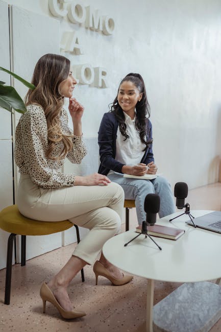 Positive multiracial female colleagues sitting on chairs near table with microphones and laptop while recording audio program in modern studio